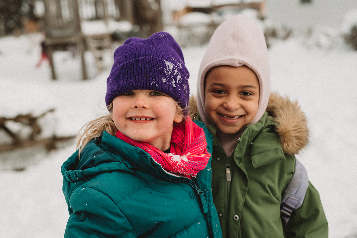 two smiling girls wearing winter hats and coats in snowy outdoors look directly into the camera