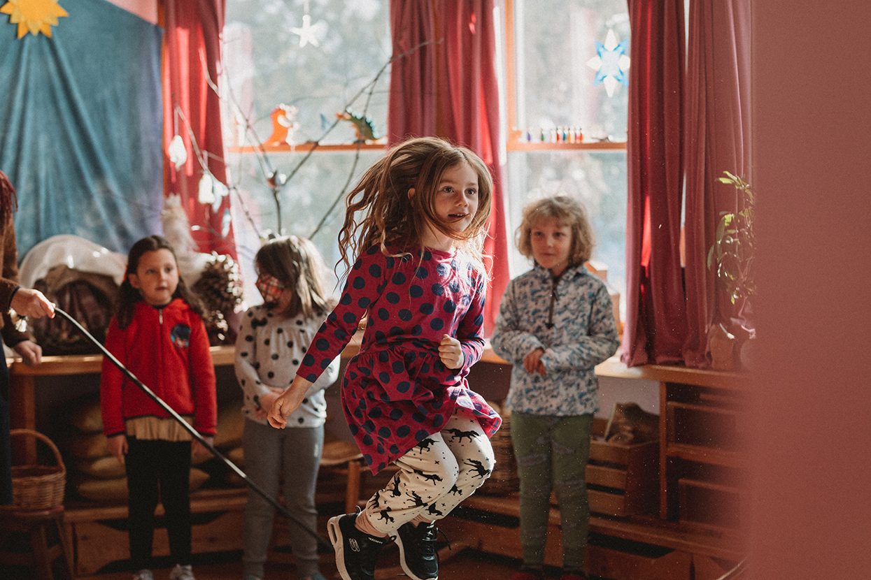 Young girl with long hair in the midst of jumping rope in an early childhood classroom; classmates observe in the background; light-filled room with lots of windows