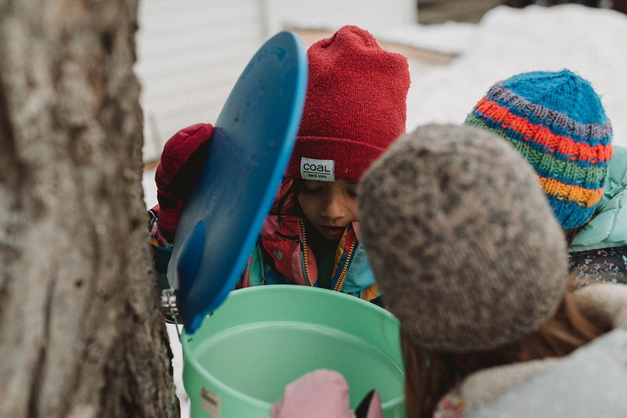 Three young children wearing winter gear peer into sap bucket hanging off a tree outdoors in the winter.