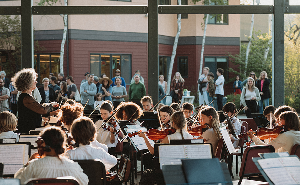Students performing with their string instruments under the open air pavilion; Strings and orchestra ensemble teacher Alison Eldredge conducts