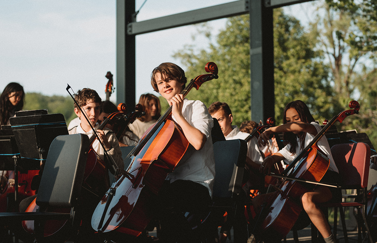 Middle school strings students ready to perform under school's outdoor pavilion. A male student with a bass is looking directly into the camera and smiling. The scene is sunlit.