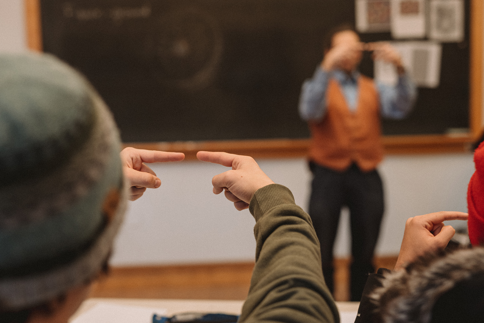 Student in foreground holding two index fingers pointing toward one another in front of their face; teacher in background demonstrating the same action