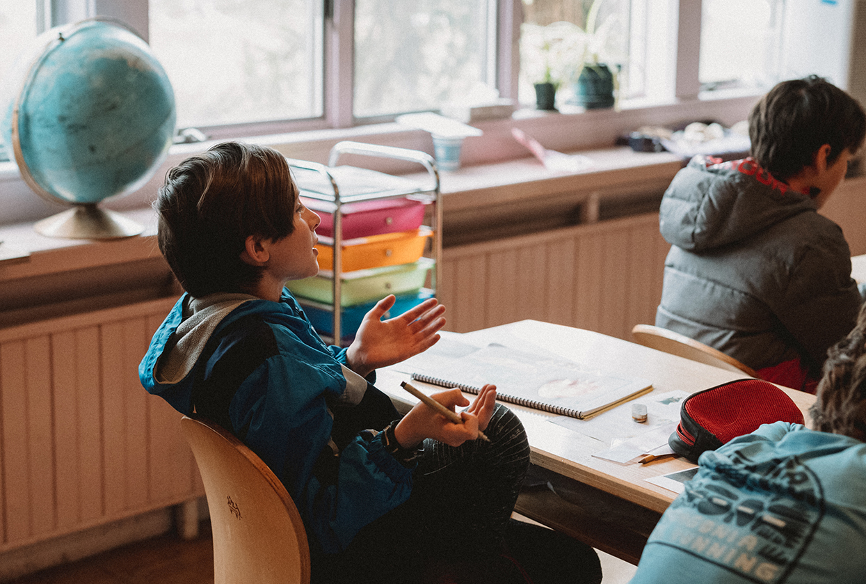 Middle school student in classroom engaged in class discussion with gesturing hands. It is a light-filled room with windows in the background