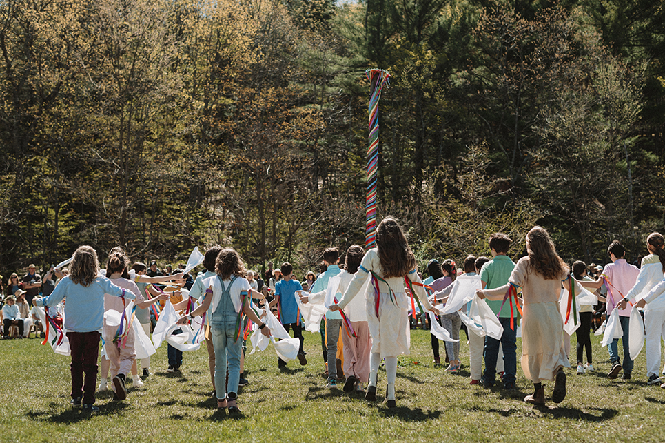 Group of children perform a May Day dance around the May pole; they have colorful ribbons tied around their upper arms and are carrying white flags