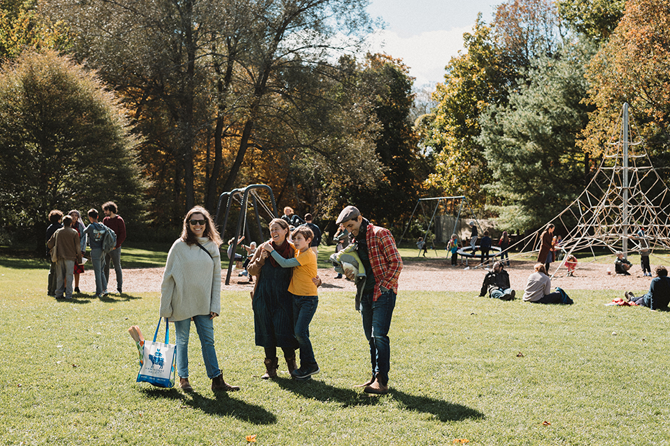 A group of happy adults and one child socializing near the school playground; they are laughing, and one of the women is holding a Hawthorne Valley shopping bag with a baguette sticking out of it; it is a sunny, early Fall day