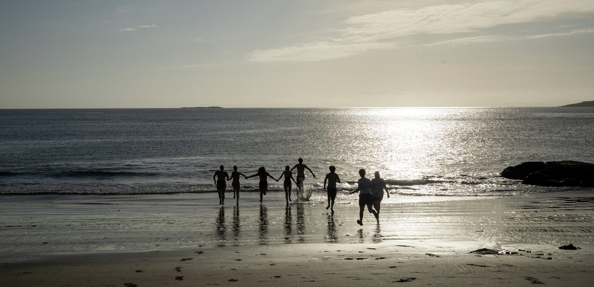 a line of high school students silhouetted at the ocean; they are running toward the water together at sunset; the beach and water are both silvery in color