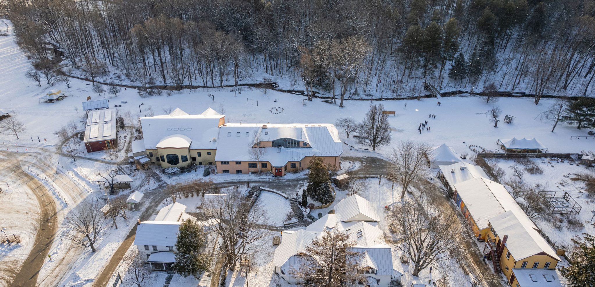 Aerial view of school campus in winter; creek can be seen just inside the tree line. From top to bottom of the photo it shows the baren woods, the thin creek, and all of the campus buildings including the middle school, early childhood, Turose, and assembly hall.