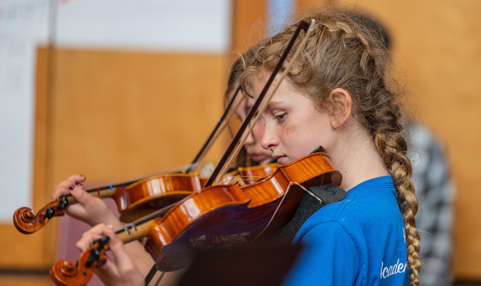 two girls playing violins sitting in a row; student in foreground wears a long braid and blue shirt
