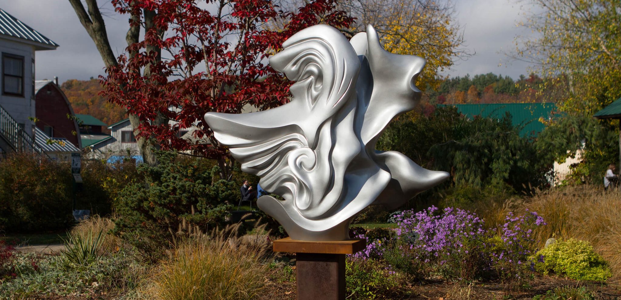 The sculpture, "Flight" is located in a garden bed just across the street from the school. It is pictured here in autumn surrounded by plant life like bushes, wildflowers, and trees.