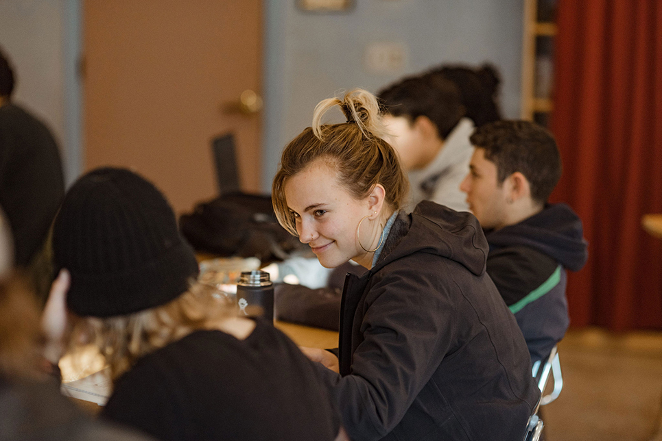 Students seated in a high school classroom; a young woman wearing her hair up in a clip is smiling and engaged in conversation with another student next to her