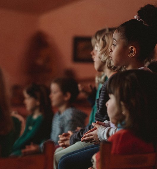 Group of kindergarten students watching a puppet performance. The focus is on the left side of three young girls with other blurry students in the background.