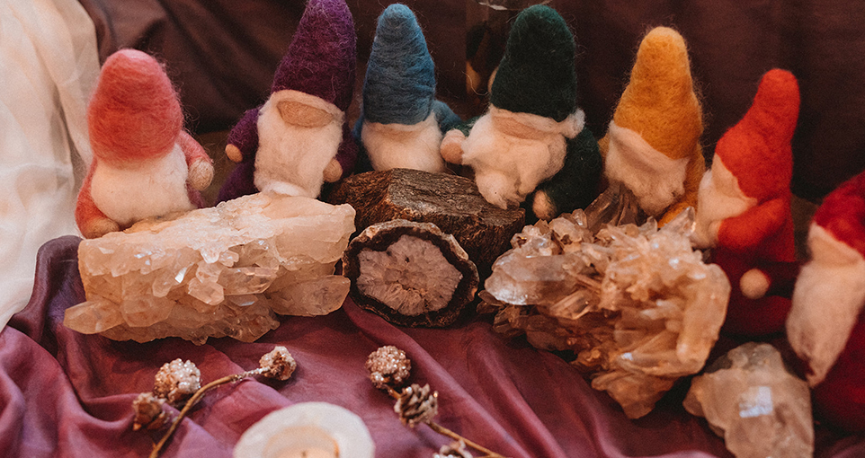 Row of colorful peg doll gnomes on a mauve-colored cloth; each is wearing a different colored felt hat and there are crystals on the table as well as pinecones
