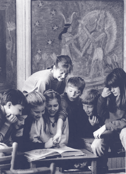 Historical photo featuring group of seven students gathered around a desk all looking at a book that is open on the desk; one student points to something on the page; there is a chalkboard drawing in the background featuring Egyptian figures
