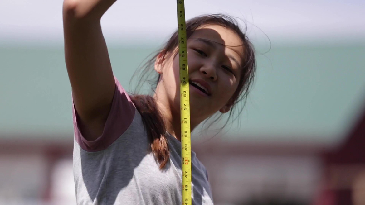 Young Asian woman working with a tape measure outdoors during a surveying block; it is a close up image; one hand is over her head out of range of the image, and the tape measure is vertical from top to bottom of the photo; it is sunny and there is a shadow across her face