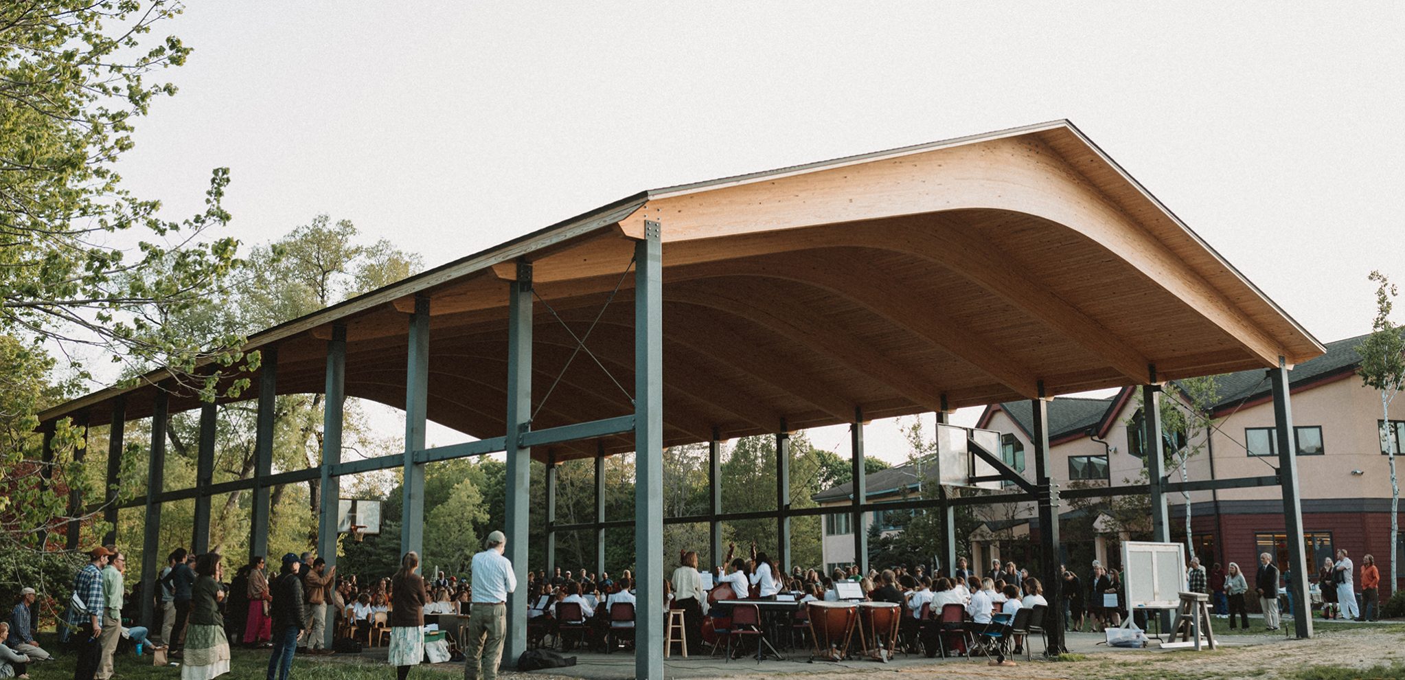 View of school open air pavilion during a concert from an angle. The children are in the distance under the pavilion and the middle school is in the background behind the pavilion.