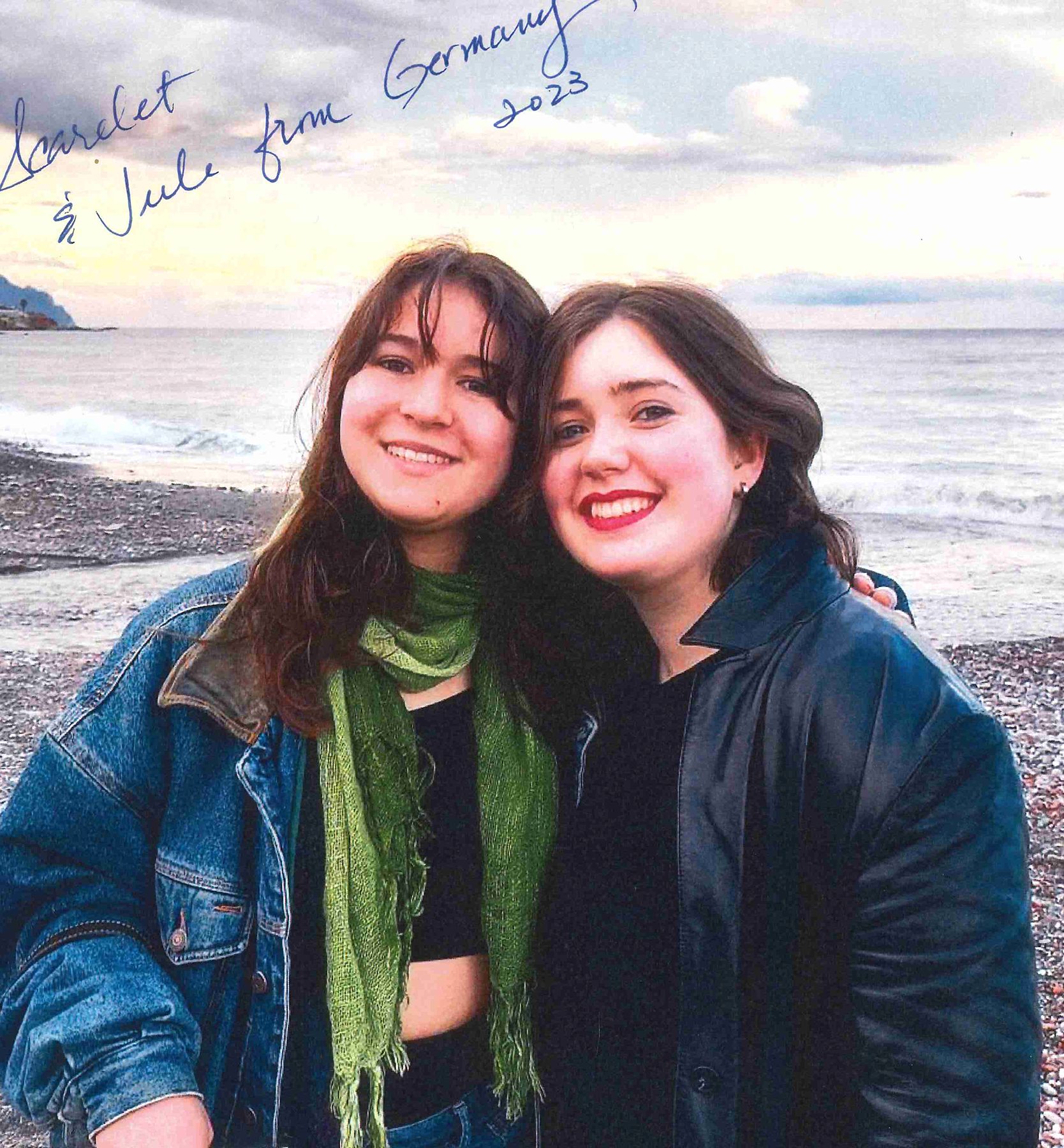 Two female students stand close together with their heads touching on a beach; they are both smiling and looking into the camera; the woman on the left is wearing a jean jacket and a green scarf; the woman on the right is wearing a black leather jacket and black shirt; both have shoulder-length brown hair; there is handwriting on the photo that reads, "Scarlet & Jule from Germany 2023"