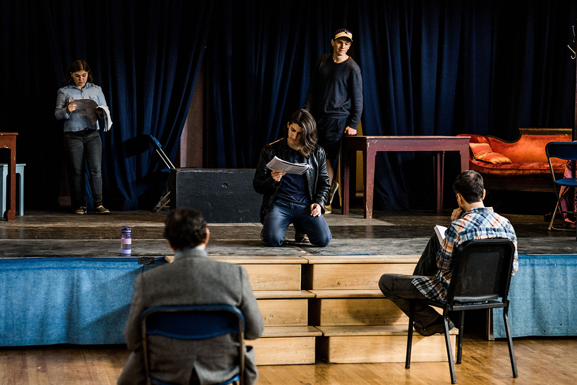 Students on stage during a play rehearsal; one male student is on his knees and holding a script; there is a female student and a male student on stage standing behind him, and two teachers seated off stage