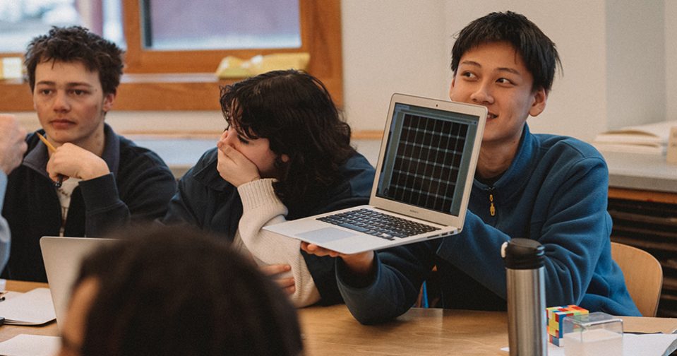 Three high school students - two males and one female - at a table engaged in a lesson; one student is holding a tablet and showing the screen to someone off camera. The other students sit to the right of him, one covering her mouth, laughing, the other staring ahead.