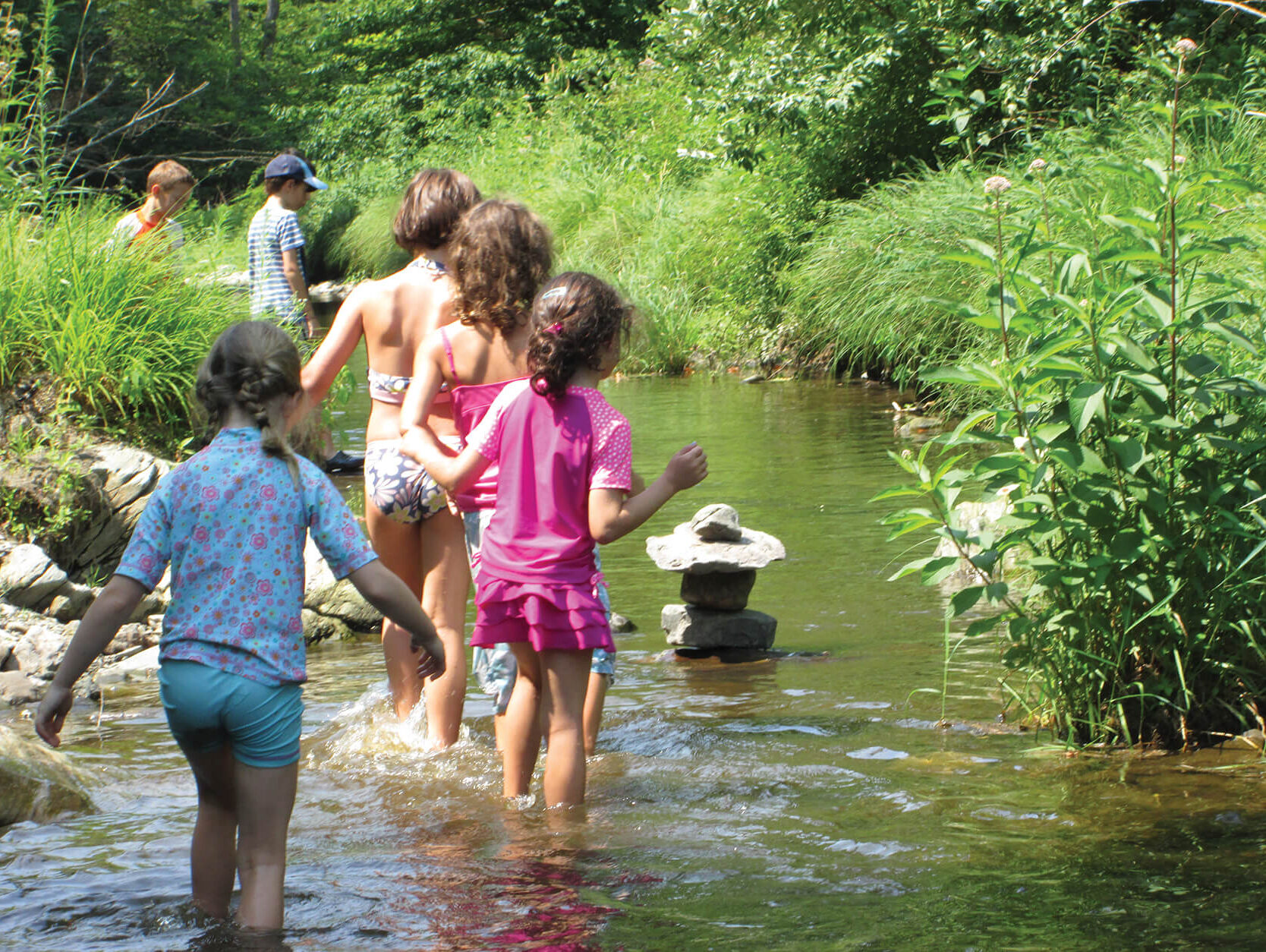 Young children walking in stream during summer; all are wearing shorts and swim suits; there is a stack of rocks in the middle of the stream