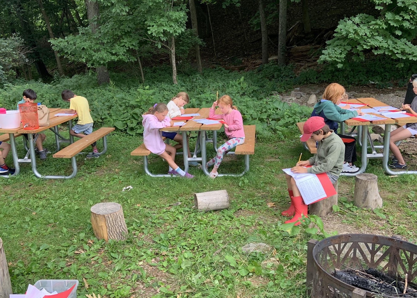 Children from Hickory Program Day Camp sitting on picnic tables outside writing on paper